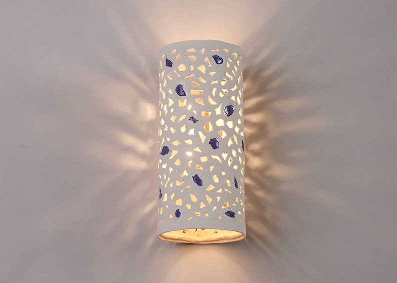 May include: A white ceramic wall sconce with a cylindrical shape. The sconce features a cut-out design with blue accents. The light shines through the cut-outs, creating a pattern on the wall. The sconce is mounted on a white wall.