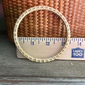 May include: A circular beige woven rattan bangle positioned in front of a wooden ruler. The ruler shows the bangle has an outer diameter of approximately 3.5 inches. The bangle features a consistent, repeating woven pattern throughout its structure.