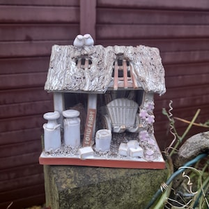 Könnte beinhalten: Eine Keramik-Häuschen-Figur mit Strohdach, einem kleinen Wagen und Milchkannen. Das Häuschen ist weiß mit braunen Akzenten bemalt und trägt ein Schild mit der Aufschrift "Pond Farm".