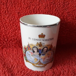 Puede incluir: Taza de cerámica blanca con un borde plateado. La taza presenta una ilustración colorida del rey Jorge V y la reina María, con el texto "In Commemoration 1910 - 1935 Silver Jubilee".