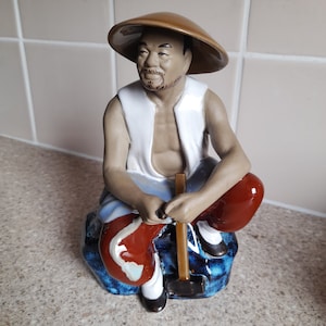 May include: A ceramic figurine of a man wearing a brown conical hat and red and blue clothing, sitting on a blue and white rock with a wooden mallet in his hand.