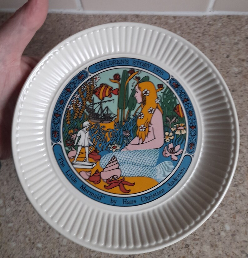 May include: A white ceramic plate with a blue border and a colorful illustration of a mermaid and a ship. The plate reads "Children's Story 1975" and "The Little Mermaid" by Hans Christian Andersen.