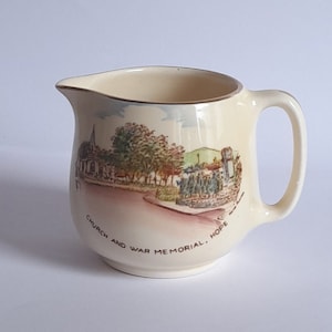 Puede incluir: Una jarra de cerámica de color crema con asa. La jarra tiene una escena de una iglesia y un monumento de guerra con el texto "Church and War Memorial, Hope" en la parte inferior.
