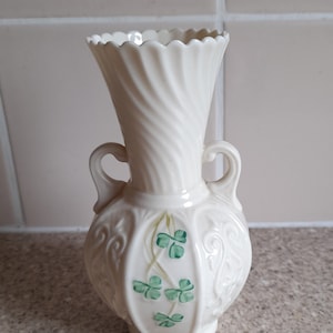 May include: Cream-colored ceramic vase with a fluted rim and two small handles. The vase features a swirling design on the neck and decorative shamrock motifs in green. A decorative piece for home decor.