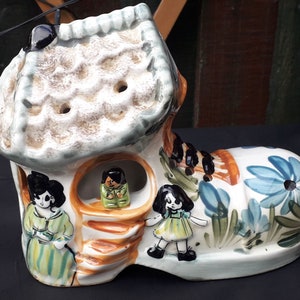 Puede incluir: Una casa de cerámica con forma de zapato con un techo verde y paredes blancas. La casa tiene una pequeña puerta y ventanas, y hay tres pequeñas figuras en el interior. El zapato está decorado con flores y hojas en tonos azules, verdes y amarillos.