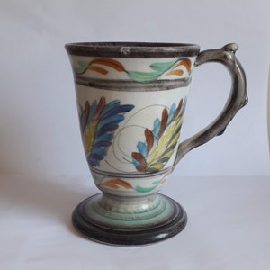 May include: A white ceramic mug with a footed base and a brown handle. The mug is decorated with colorful floral designs in shades of blue, green, yellow, orange, and brown.