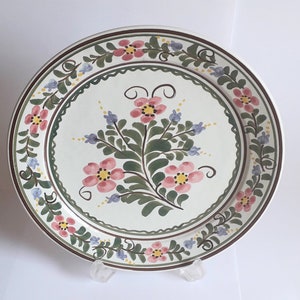 Może przedstawiać: Biały ceramiczny talerz z kwiatowym wzorem. Talerz ma zieloną obwódkę z różowymi i niebieskimi kwiatami. Środek talerza zdobi większy różowy kwiat z zielonymi liśćmi.