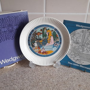 May include: A white ceramic plate with a blue and yellow illustration of a mermaid. The plate is part of a Wedgwood Children's Story collection from 1975. The plate is displayed on a stand with a blue and white box.