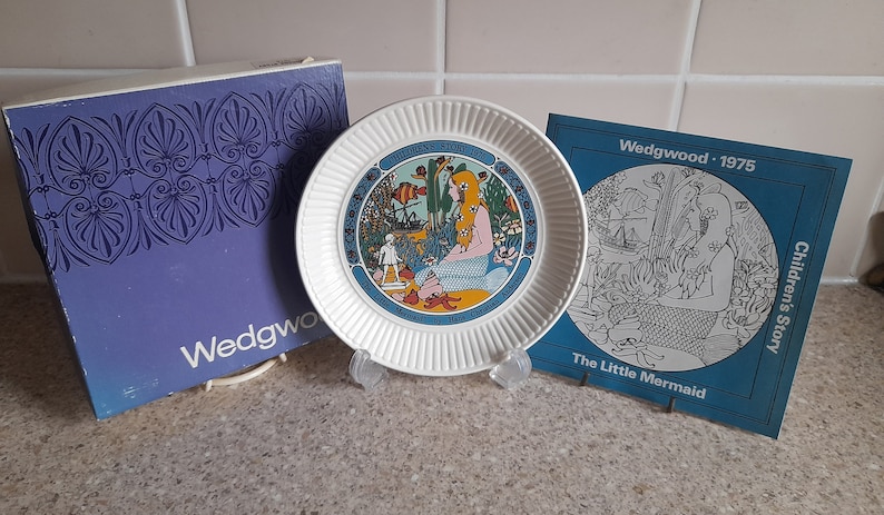 May include: A white ceramic plate with a blue and yellow illustration of a mermaid. The plate is part of a Wedgwood Children's Story collection from 1975. The plate is displayed on a stand with a blue and white box.