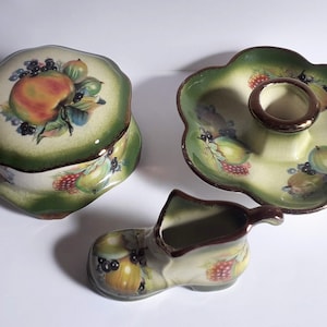 May include: A set of three ceramic trinket dishes with a green and white floral design featuring fruit and berries. The dishes are shaped like a round box, a shallow dish with a candle holder, and a shoe.
