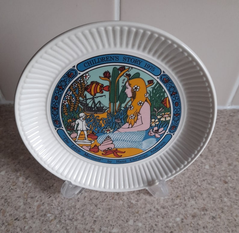May include: A white ceramic plate with a blue and yellow illustration of a mermaid and a ship. The plate has the text "Children's Story 1975" and "The Little Mermaid by Hans Christian Andersen" around the illustration.