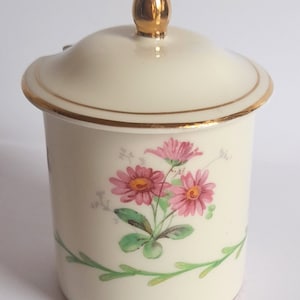 May include: A white ceramic jar with a gold lid and floral design. The jar has a pink and green floral pattern with a gold trim around the top and bottom.