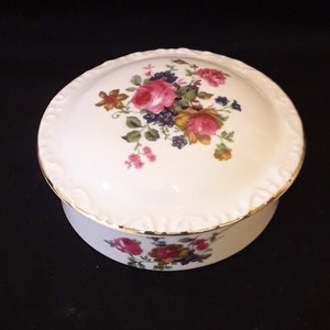 Könnte beinhalten: Eine weiße Porzellan-Schmuckdose mit Blumenmuster. Der Deckel zeigt eine Ansammlung von rosa, roten und gelben Rosen mit grünen Blättern und blauen Blüten. Die Dose hat einen goldenen Rand.