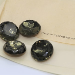 May include: Four dark green glass buttons with faceted edges. The buttons are on a white surface with the text "MADE IN CZECHOSLOVAKIA" printed on it.