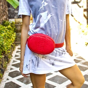 May include: A red snakeskin fanny pack worn around the waist of a person wearing a white t-shirt with a grey design.