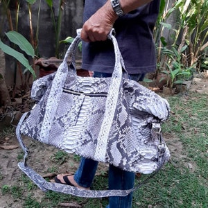 Puede incluir: Una bolsa de viaje de piel de serpiente blanca y gris con una correa larga. La bolsa tiene una cremallera y un asa superior.