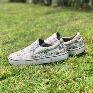 Op de afbeelding: Een paar witte slip-on schoenen met een slangenhuidpatroon. De schoenen hebben een witte rubberen zool en een zwarte rand.