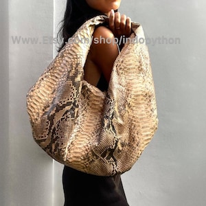 May include: A large, light brown python skin handbag with a slouchy, hobo style. The bag has a long strap and is worn over the shoulder.