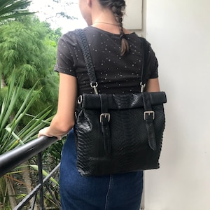 Könnte beinhalten: Schwarzer Lederrucksack mit Schlangenlederstruktur, Roll-Top-Design und zwei Schnallenverschlüssen. Die Tasche hat verstellbare Träger und silberfarbene Hardware. Die Person trägt ein dunkles T-Shirt und einen Jeansrock.