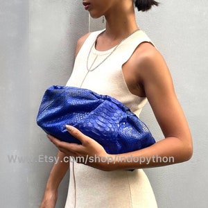 May include: A bright blue python skin clutch purse with a gathered, pleated design. The purse is held by a person wearing a light beige sleeveless dress.
