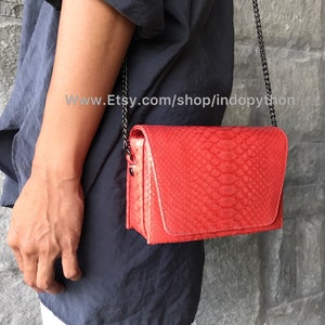 Könnte beinhalten: Eine rote Schlangenleder-Umhängetasche mit einer silbernen Kettenkette. Die Tasche hat einen Klappenverschluss und ein strukturiertes Schlangenledermuster.