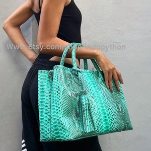 May include: A large, turquoise tote bag with a snakeskin pattern and a tassel. The bag has two handles and a shoulder strap. The bag is held by a person wearing a black tank top and black pants.