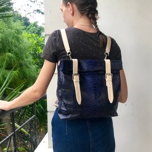 Könnte beinhalten: Ein blauer Schlangenleder-Rucksack mit cremefarbenen Riemen und Schnallen. Der Rucksack wird von einer Person getragen, die ein schwarzes Shirt und einen blauen Jeansrock trägt.