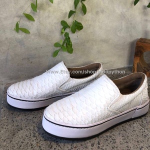 May include: White leather slip-on sneakers with a snakeskin pattern. The shoes have a white rubber sole and a black trim.