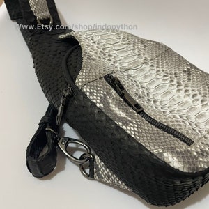 Może przedstawiać: Czarno-biała torba crossbody z wzorem skóry węża z zapięciem na zamek błyskawiczny. Torba ma czarny pasek i wykończenie, ze srebrnym metalowym zapięciem i okuciami. Projekt torby łączy tekstury i wzory.