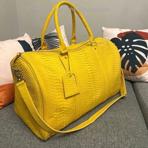 Puede incluir: Un bolso de viaje amarillo brillante con un patrón de piel de serpiente texturizado. Tiene dos asas superiores y una correa para el hombro desmontable. Una pequeña etiqueta rectangular cuelga del asa. El bolso está sobre una superficie gris.