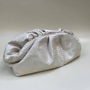 May include: A silver clutch bag with a snakeskin pattern. The bag has a gathered design and a soft, rounded shape. The material has a subtle sheen, reflecting light.
