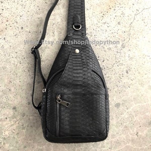 Könnte beinhalten: Eine schwarze Leder-Umhängetasche mit Schlangenhautmuster. Die Tasche hat eine Reißverschlusstasche auf der Vorderseite und einen verstellbaren Riemen für eine bequeme Passform.