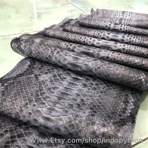 May include: A stack of grey python leather hides with a scaly texture. The hides are arranged in a fan shape, with the largest hide at the bottom and the smallest hide at the top. The hides are all of the same colour and texture. The text "www.Etsy.com/shop/indopython" is visible at the bottom of the image.