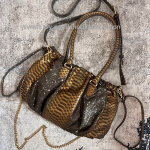 Puede incluir: Un bolso de mano de piel de serpiente marrón y dorado con una correa de cadena y una correa larga marrón. El bolso tiene un diseño fruncido y una cremallera superior.