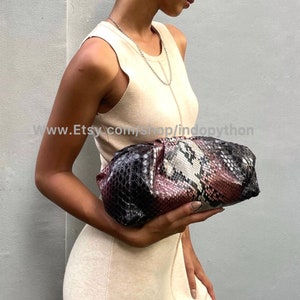 May include: A woman holds a clutch purse with a snakeskin pattern in shades of black, pink, and white. The purse has a gathered, cloud-like shape.
