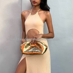 Puede incluir: Una mujer lleva un vestido halter de color melocotón claro con un detalle de corte y sostiene un bolso de mano de piel de serpiente dorada. El bolso tiene una superficie texturizada y una forma ligeramente redondeada.