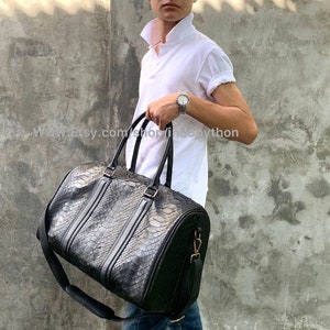 Puede incluir: Una bolsa de viaje de cuero negro con un patrón de piel de serpiente. La bolsa tiene dos asas superiores y una correa de hombro larga ajustable.