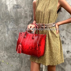 Könnte beinhalten: Eine Frau trägt ein goldenes, ärmelloses Kleid mit einem Schlangenledergürtel und eine rote Schlangenlederhandtasche mit einem Quastenanhänger.