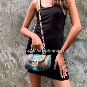 May include: A small, teal and brown snakeskin handbag with a gold chain strap and a gold lock. The bag is worn by a person wearing a black tank top.