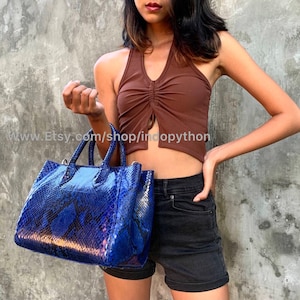 Puede incluir: Un bolso de mano de piel de pitón azul con asa superior y dos correas para el hombro. El bolso tiene un patrón texturizado de piel de serpiente.
