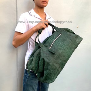 Puede incluir: Una bolsa de viaje de cuero verde oscuro con cierre de cremallera y un tirador de cremallera plateado. La bolsa está hecha de piel de pitón y tiene una superficie texturizada.