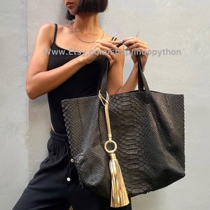 Könnte beinhalten: Eine große schwarze Ledertasche mit Schlangenhautmuster und einem goldenen Quastenanhänger. Die Tasche hat eine große Öffnung und zwei obere Henkel. Die Tasche wird von einer Person getragen, die ein schwarzes Tanktop und eine schwarze Hose trägt.