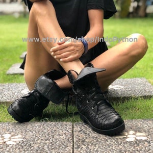 May include: Black leather boots with a snakeskin pattern and laces. The boots have a fold-over flap at the ankle and are worn by a person sitting on a grassy surface. The text "www.etsy.com/shop/IndoPython" is visible on the ground.