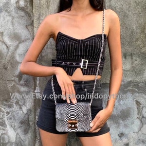 Może przedstawiać: Czarno-biała torebka ze skóry węża z srebrnym łańcuszkiem. Torebka ma złoty zamek i jest noszona przez osobę ubraną w czarno-białą, paskowaną bluzkę i czarne spodenki.