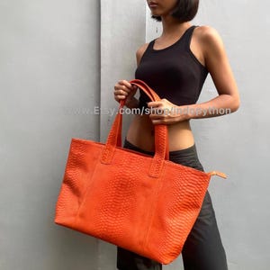 Op de afbeelding: Een feloranje tote bag met een reptielhuidtextuur. De tas heeft twee handvatten en een rechthoekige vorm. De tas wordt door een persoon vastgehouden. De tas heeft een levendige oranje tint.