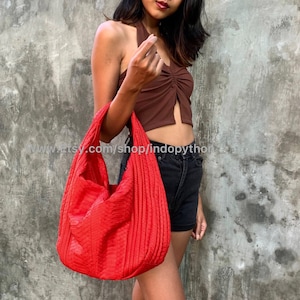 May include: A red quilted tote bag with a textured surface. The bag is being held by a person wearing a brown halter top and black shorts.