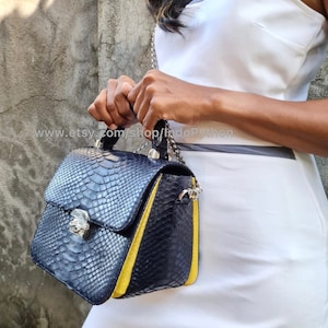 Puede incluir: Un pequeño bolso de mano azul oscuro con ribete amarillo y una correa de cadena plateada. El bolso está hecho de piel de serpiente y tiene un cierre de solapa con un broche plateado.