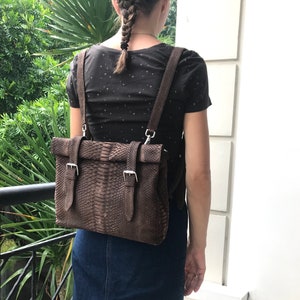 Może przedstawiać: Brązowy skórzany plecak z wzorem skóry węża. Torba ma regulowane paski i srebrne klamry. Plecak jest noszony na ramionach. Osoba ma na sobie czarną koszulę i dżinsową spódnicę.