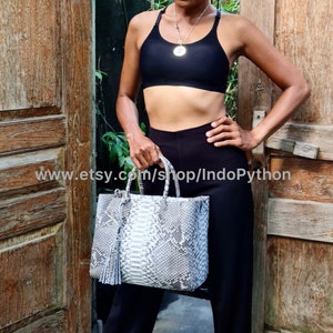 Może przedstawiać: Biała torba typu tote ze skóry pytona z frędzlem. Torba ma górny uchwyt i jest wykonana z prawdziwej skóry pytona.