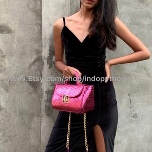 Op de afbeelding: Een felroze handtas van slangenleer met een gouden kettingband en een gouden slot. De tas is klein en heeft een vierkante vorm.